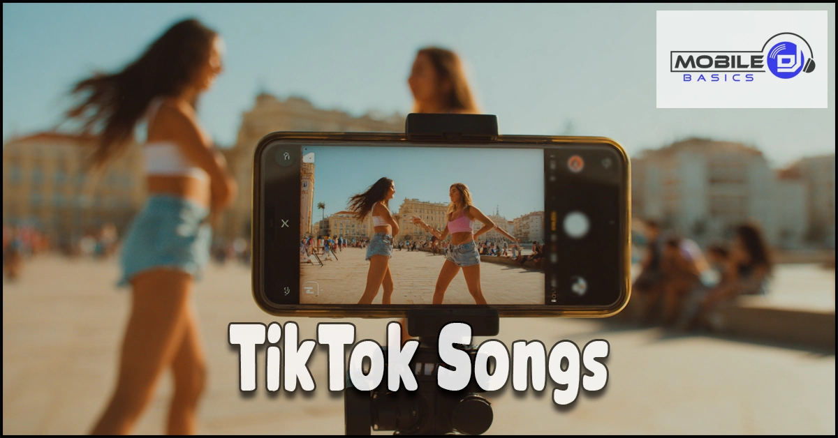 Two women dance outdoors, filmed by a smartphone on a tripod. The phone screen shows them moving to popular TikTok Songs. Text reads “TikTok Songs.” The Mobile Basics logo appears in the top right corner.