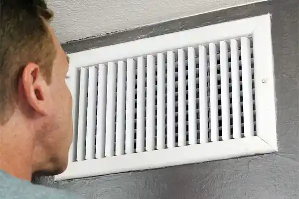 A Moore Home Services technician carefully inspects a spotless white air vent on a gray wall, ensuring your HVAC system is running efficiently. Trust our clean, professional team to deliver expert heating, cooling, plumbing, and drain solutions—backed by our satisfaction guarantee for your peace of mind.