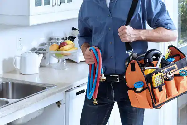 A friendly Moore Home Services technician in a crisp blue uniform stands beside a spotless kitchen sink, ready to tackle your plumbing needs with a well-stocked tool bag and high-quality hoses. Count on us for expert plumbing solutions, clean service you can trust, and our satisfaction guarantee every time.