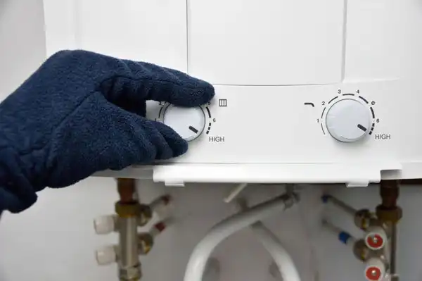 A Moore Home Services technician, wearing protective gloves, expertly adjusts the temperature control on a white water heater, ensuring optimal performance. Clean, organized pipes are visible below the unit, reflecting our commitment to tidy plumbing work. You can trust our team for reliable water heater and plumbing solutions—backed by satisfaction guarantees and professional service every time.