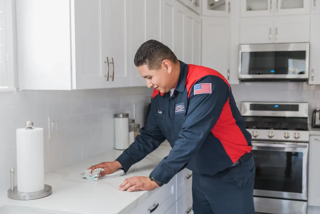 A friendly Moore Home Services technician, dressed in our signature navy and red uniform, carefully wipes down a spotless white kitchen countertop near gleaming stainless steel appliances and crisp white cabinets. Our trusted experts always provide reliable plumbing service with a commitment to leaving your home as clean as we found it—guaranteed! Choose Moore for courteous professionals who put your comfort and peace of mind first.