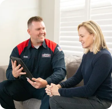 A friendly Moore Home Services technician, dressed in a clean uniform, consults with a homeowner inside her residence. Both are smiling as the technician explains HVAC, plumbing, or drain service options on a tablet—delivering trusted solutions and exceptional care you can count on. Our team guarantees your satisfaction and always leaves your home as spotless as we found it.