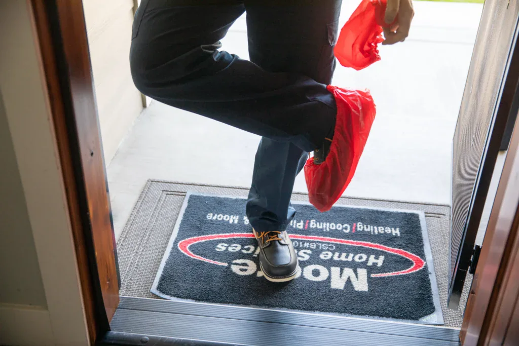 A Moore Home Services pro puts on red shoe covers at your door—ready to deliver HVAC, plumbing, or drain help with total care.