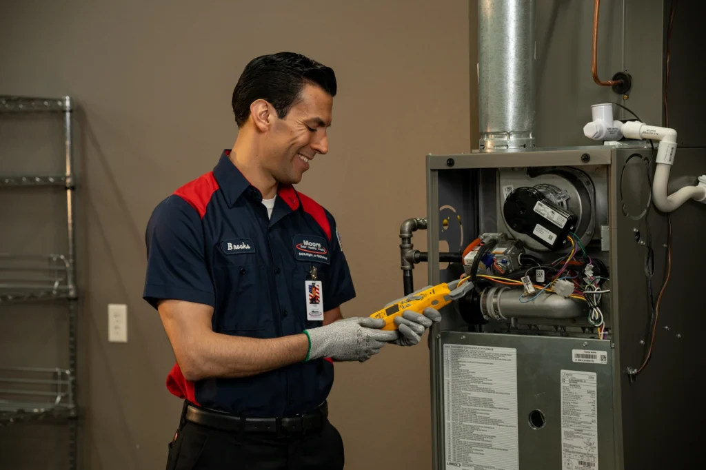 One of our friendly, uniformed Moore Home Services technicians carefully inspects the wiring inside an open furnace unit using a specialized handheld device in a homeowner’s living room. We pride ourselves on expert HVAC service, keeping your home comfortable and safe with our trustworthy professionals, backed by guarantees and a commitment to leaving your home as clean as we found it.