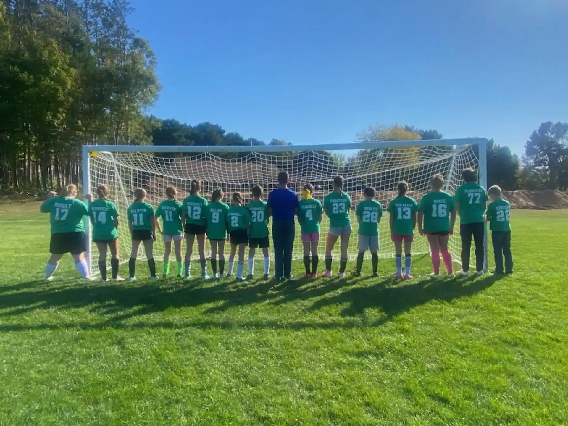 Group of children in soccer uniforms standing in front of a goal on a grassy field.