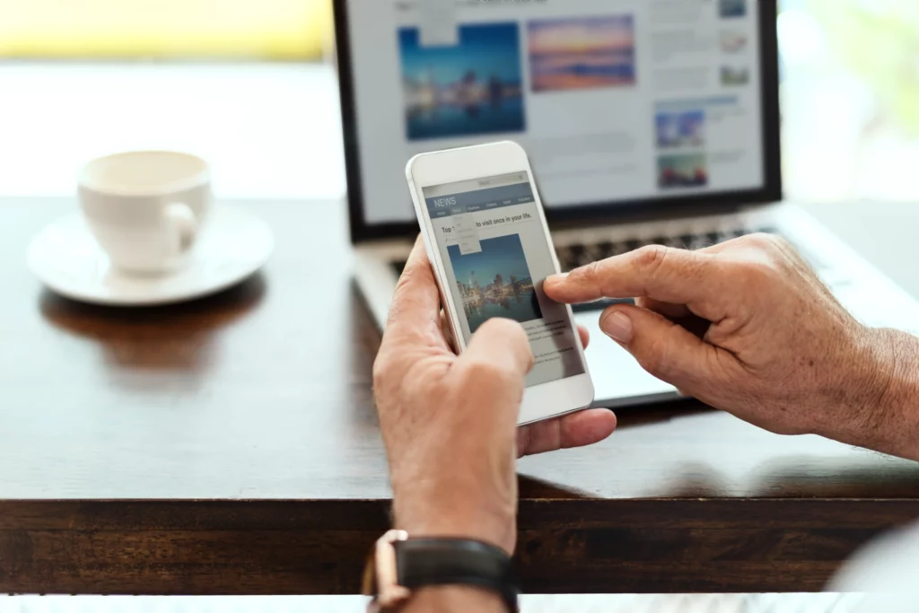 A person engages with their smartphone at a table, where an open laptop and steaming cup of coffee suggest a seamless integration of mobile-first design principles in their tech-savvy environment. mobile first web design