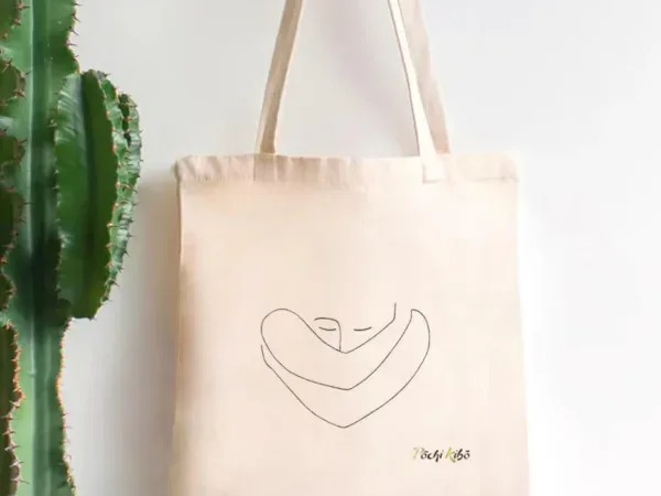 tote-bag-confiance-en-soi Tote-bag Confiance en soi accroché à un porte-manteau, avec un tracé représentant une personne qui se prend elle-même dans ses bras, et le logo Pochi Kibo en bas à droite, fond blanc avec un cactus à gauche.