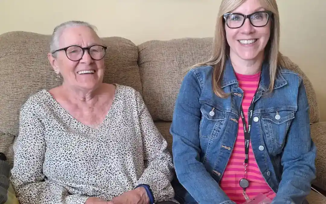 Elderly woman and caregiver smiling on a sofa in a cozy home setting.