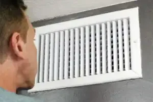 A knowledgeable technician carefully examines a white air vent on a gray wall, ensuring reliable home comfort for our community.
