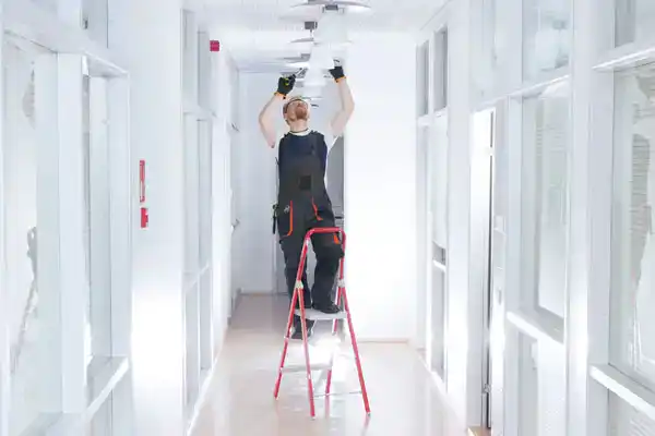 A Service Champions technician on a stepladder expertly services a ceiling light in a bright, inviting hallway with glass-paneled walls.
