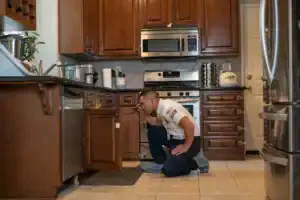 A friendly Service Champions technician in uniform inspects a kitchen cabinet with care during a plumbing repair in Brea, CA.