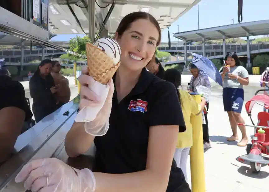 A woman wearing gloves happily holds up a complimentary ice cream cone from a Service Champions food truck in Anaheim, offering a warm smile to the camera. In the background, several community members line up under sunny skies, reflecting Service Champions’ dedication to bringing comfort and care to Southern California neighborhoods as your trusted local HVAC, plumbing, and drain experts.