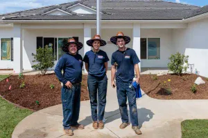 Three veterans, dressed in matching hats and navy uniforms, stand smiling together on a sidewalk in front of a well-maintained white house with a beautifully landscaped yard—a scene that reflects the trusted, dependable service and commitment to community that Service Champions brings to homes across Southern California.