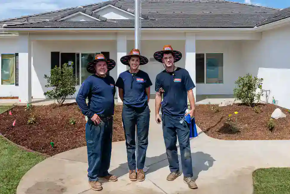Three veterans, dressed in matching hats and navy uniforms, stand smiling together on a sidewalk in front of a well-maintained white house with a beautifully landscaped yard—a scene that reflects the trusted, dependable service and commitment to community that Service Champions brings to homes across Southern California.