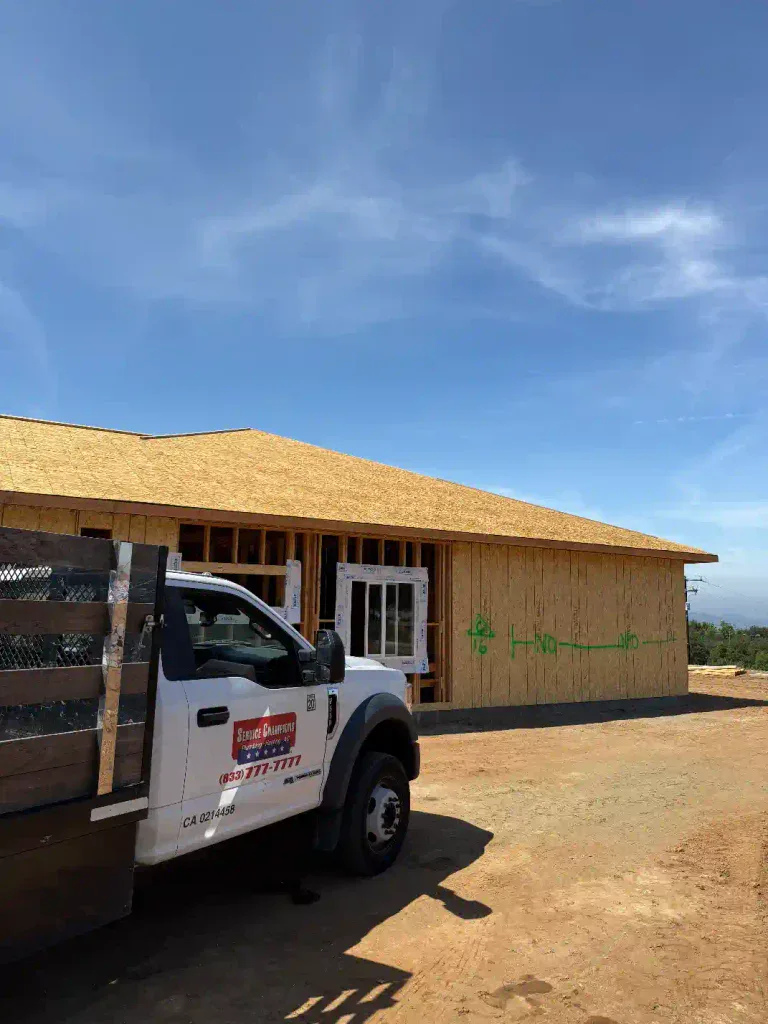 A reliable white work truck is stationed in front of a partially built home featuring sturdy wooden framing and a plywood roof, illuminated by the clear blue Southern California sky. Demonstrating Service Champions’ commitment to dependable home comfort, our team actively supports initiatives like Homes For Our Troops—proudly providing local, expert HVAC, plumbing, and drain services with the care and trust our community deserves.