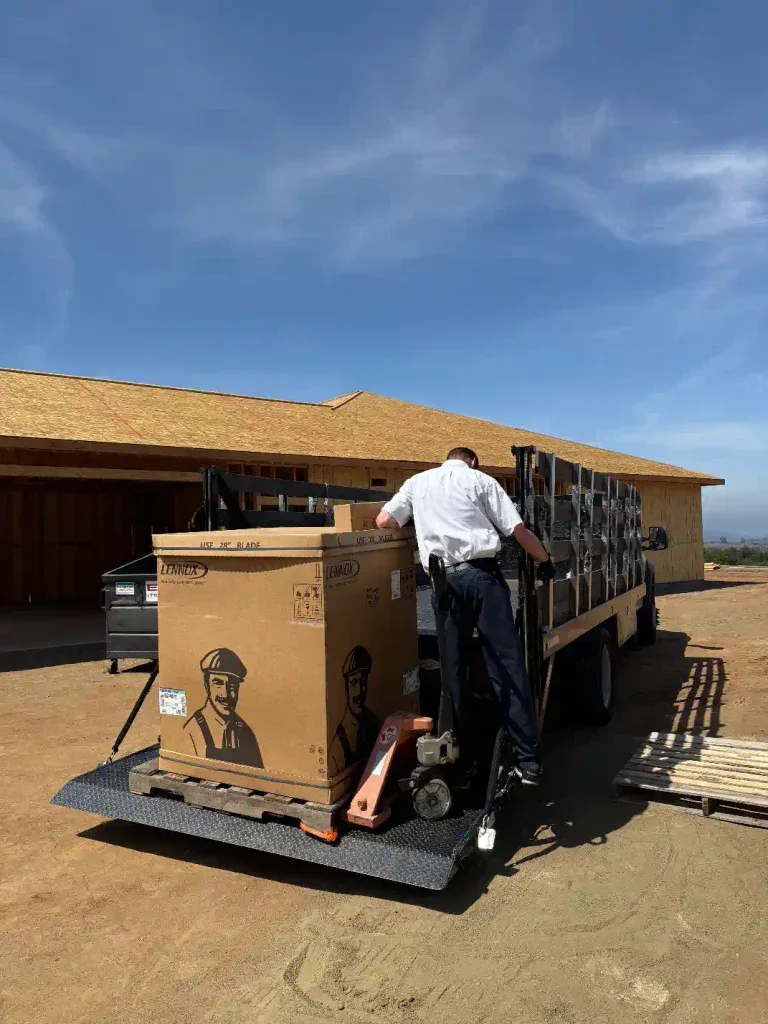 A dedicated worker carefully unloads a large cardboard box from a flatbed truck at a Homes For Our Troops construction site, beneath a clear blue sky. Their helpful spirit and commitment echo how Service Champions proudly supports Southern California communities, delivering dependable home comfort solutions with reliability and care to those who need it most.