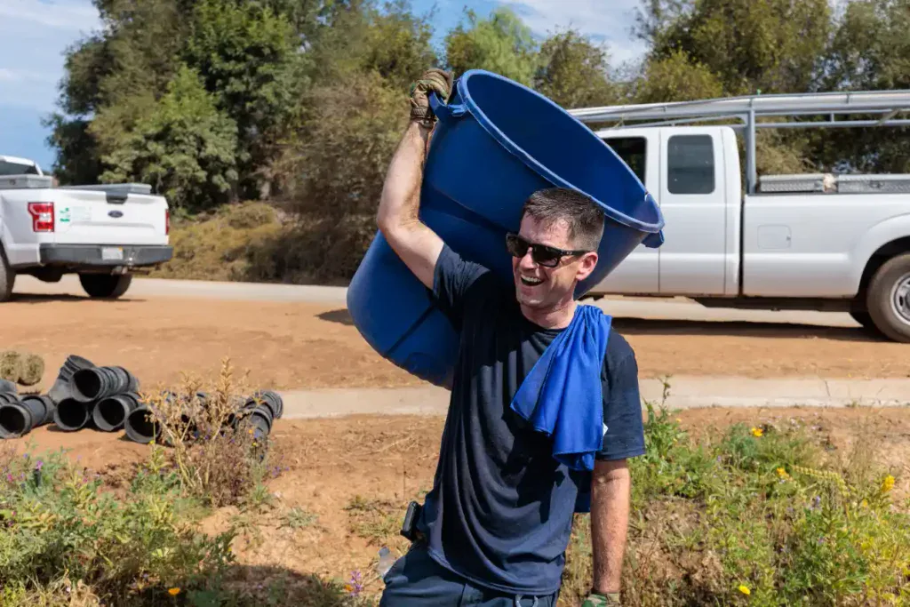 A dedicated Service Champions team member, wearing sunglasses, carries a large blue barrel on his shoulder outdoors near a white company truck. This moment captures the helpful spirit, reliability, and customer-first attitude that define Service Champions as Southern California’s trusted local expert in HVAC, plumbing, and drain services.