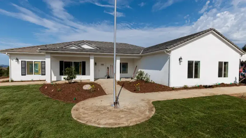A single-story white house with a gray roof, spacious windows, and a central entrance sits amid a beautifully landscaped yard with fresh green grass and a flagpole along the front walkway. This inviting home represents Service Champions’ dedication to creating comfortable, dependable living spaces for Southern California families—reflecting their customer-focused expertise in HVAC, plumbing, and drain services. As trusted local experts, Service Champions is committed to providing helpful and reliable solutions that honor the comfort needs of every home they serve.