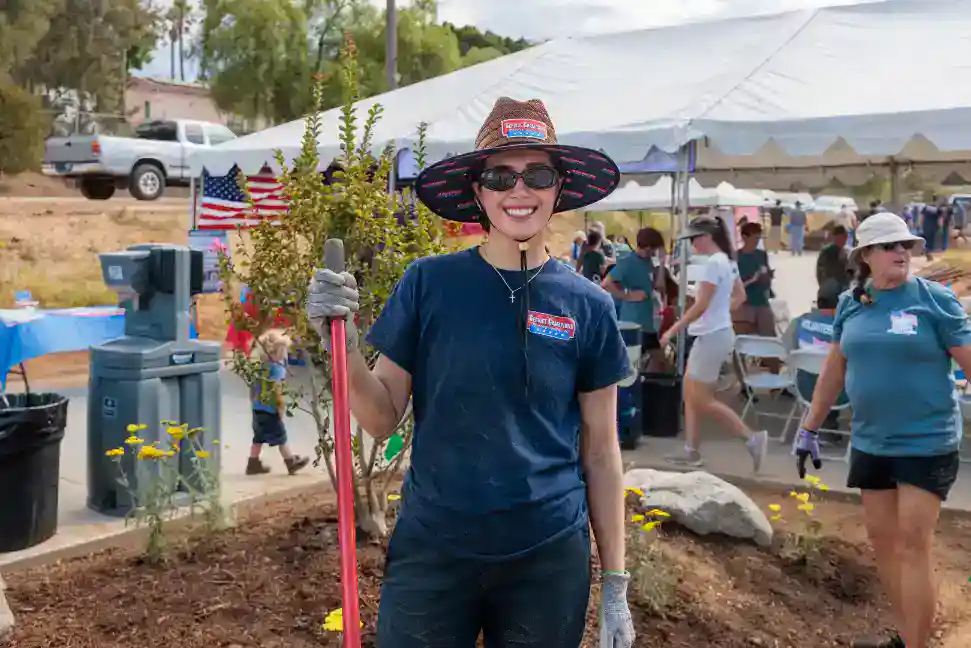 A woman wearing a hat and gloves participates in gardening at an outdoor community event, working alongside fellow Veterans. Tents and an American flag can be seen in the background, highlighting a spirit of teamwork and dedication—values that mirror Service Champions’ dependable, customer-focused approach to HVAC, plumbing, and drain services for Southern California families.