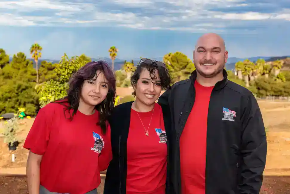 Three dedicated team members stand outdoors, two in red shirts and one in a black jacket, all displaying coordinated Homes For Our Troops logos. Set against a backdrop of palm trees and a partly cloudy Southern California sky, they reflect Service Champions’ genuine commitment to serving the community with expert HVAC, plumbing, and drain solutions—supporting both customers’ comfort and important causes with trustworthiness and care.