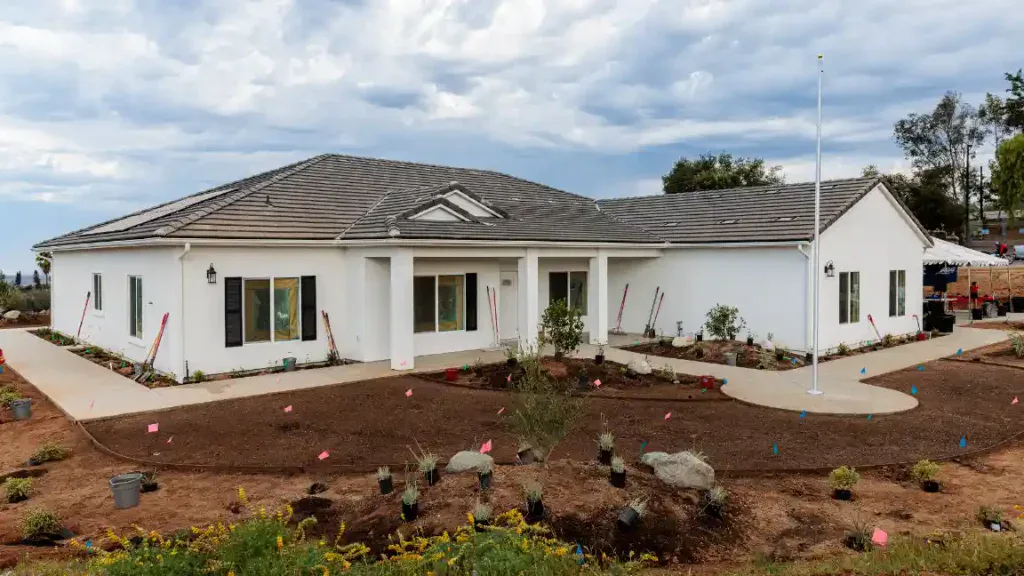 A welcoming single-story white home with a gray tiled roof, inviting covered entryway, and well-maintained front yard sits under a gentle cloudy sky. As a proud tribute to Veterans through the Homes For Our Troops initiative, this residence embodies the warmth and reliability that Service Champions brings to Southern California families—delivering expert HVAC, plumbing, and drain solutions you can trust for lasting comfort and peace of mind.