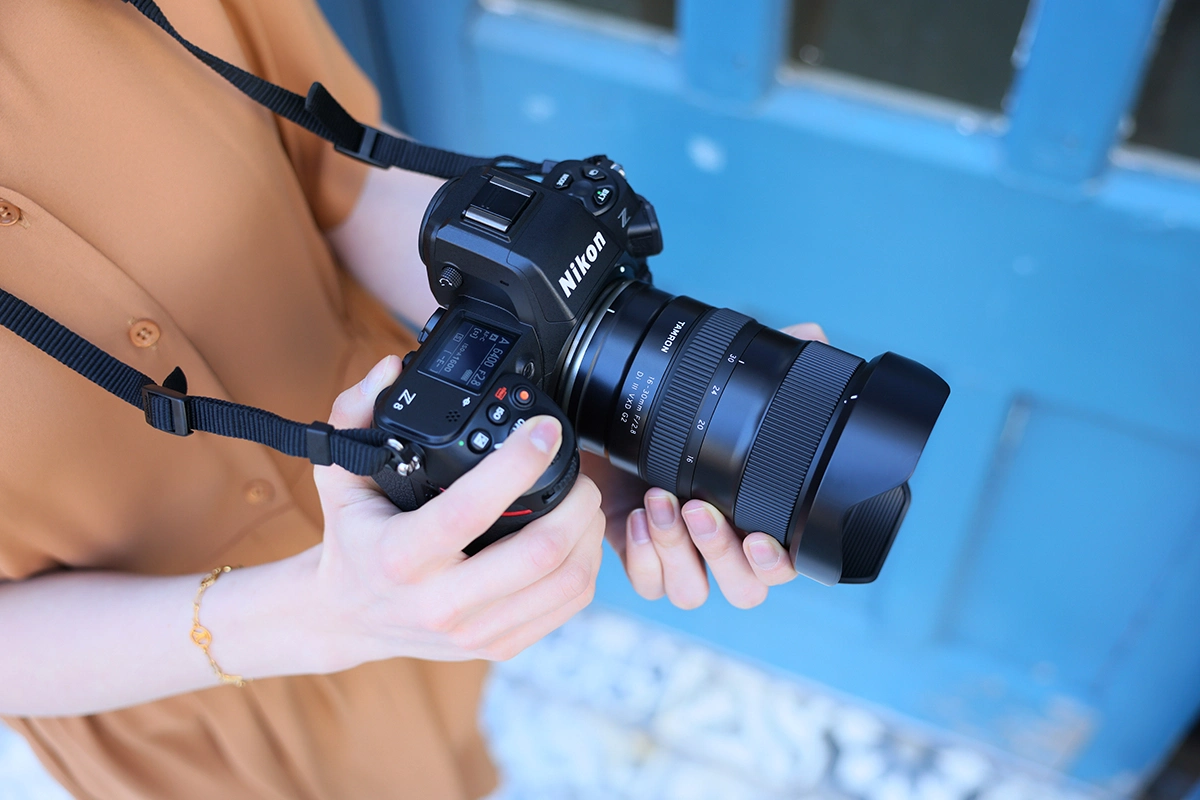 A woman in a brown shirt holding a Tamron 16-30mm lens mounted on a Nikon camera.