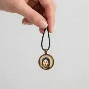 Hand holding the black cord loop to display a hanging round Jesus Christ Pantocrator icon glass pendant.