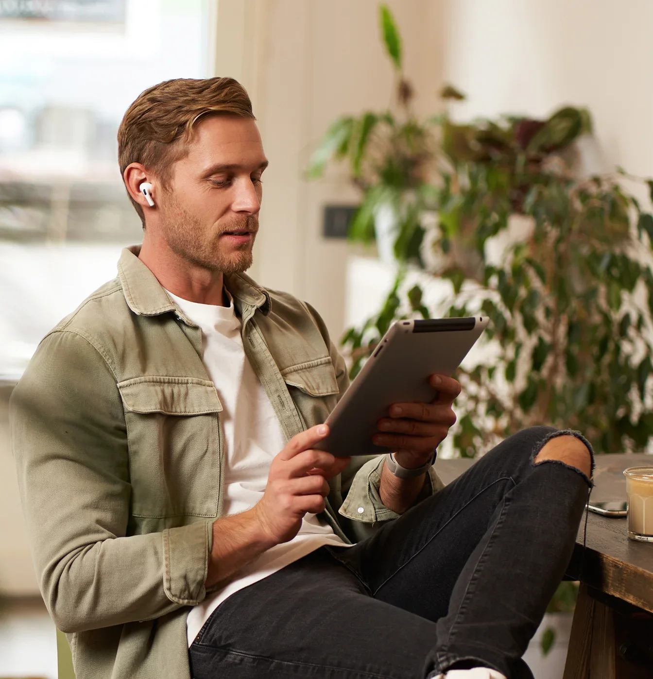 A man wearing wireless earbuds sits at a table using a digital tablet.