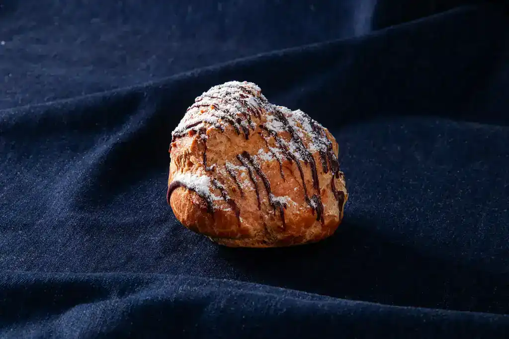 Fresh cream puff with chocolate drizzle on dark fabric background.
