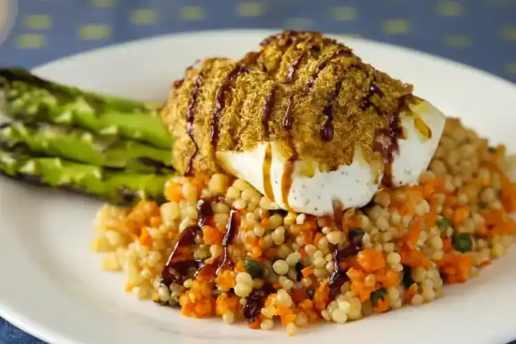 Pistachio-crusted cod with vegetables and couscous on a white plate.