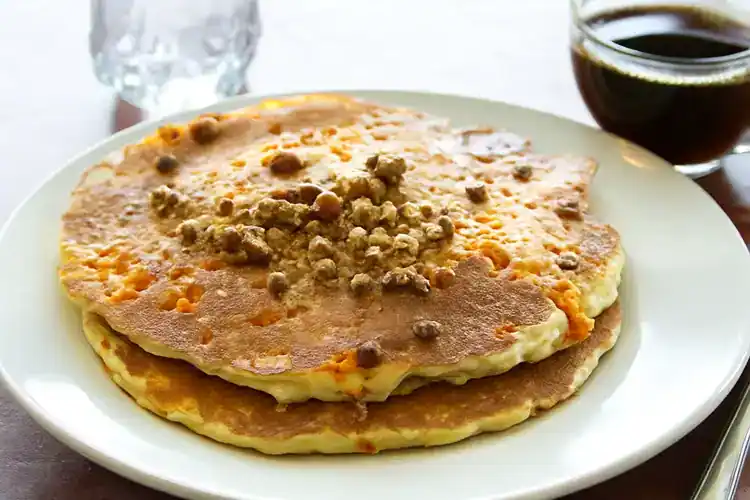 Stack of sweet potato pancakes topped with crumbled cheese or nuts, served on a white plate with a s.