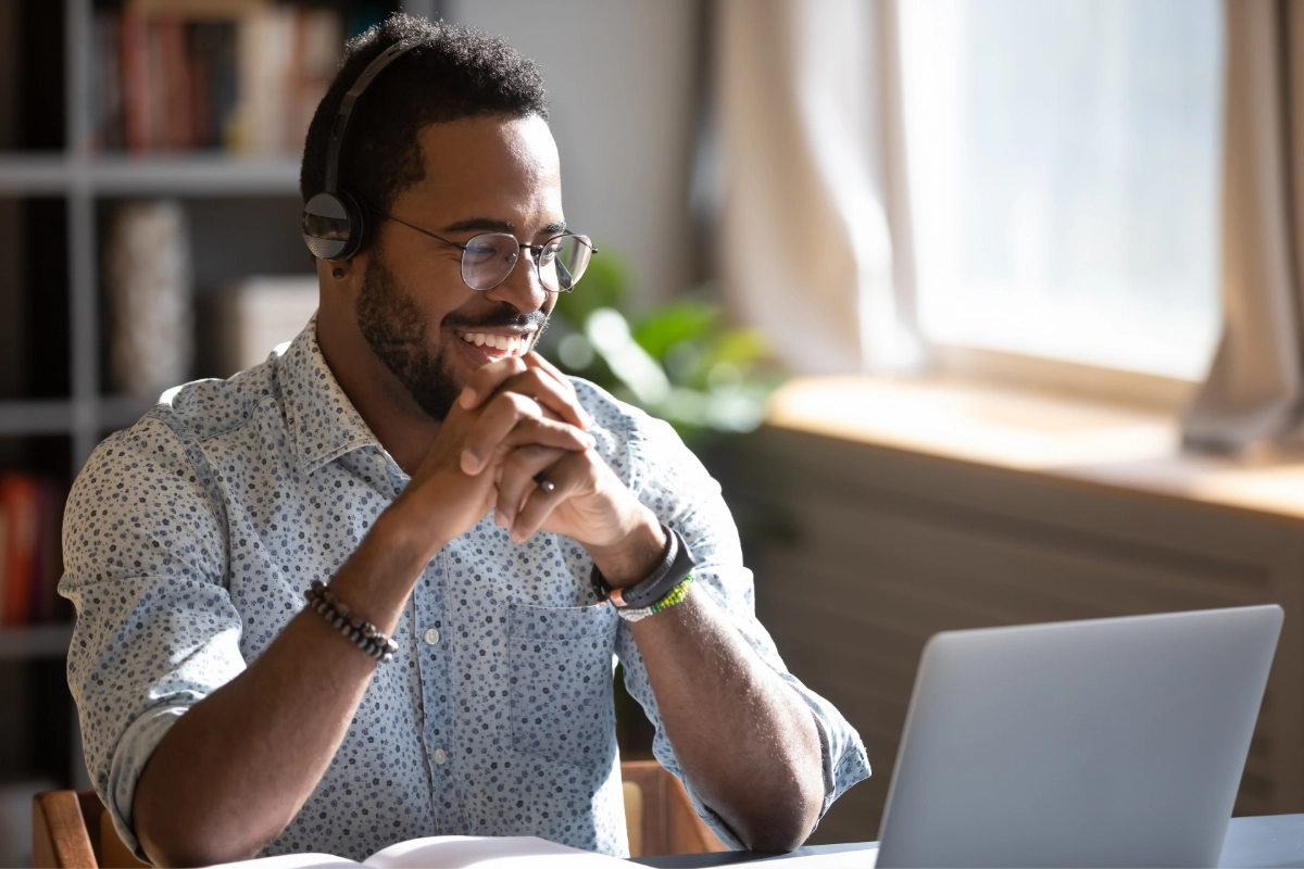 A person smiles and clasps their hands near their face while leaning their elbows on the desk and looking at a laptop.