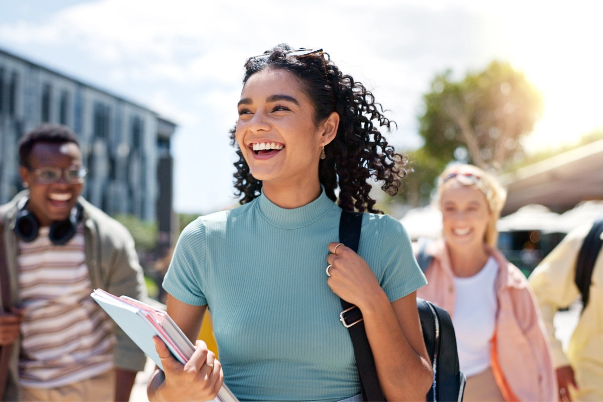 A young, smiling adult shoulders a backpack and holds a notebook in the other hand while walking with two companions outside a nearby building.
