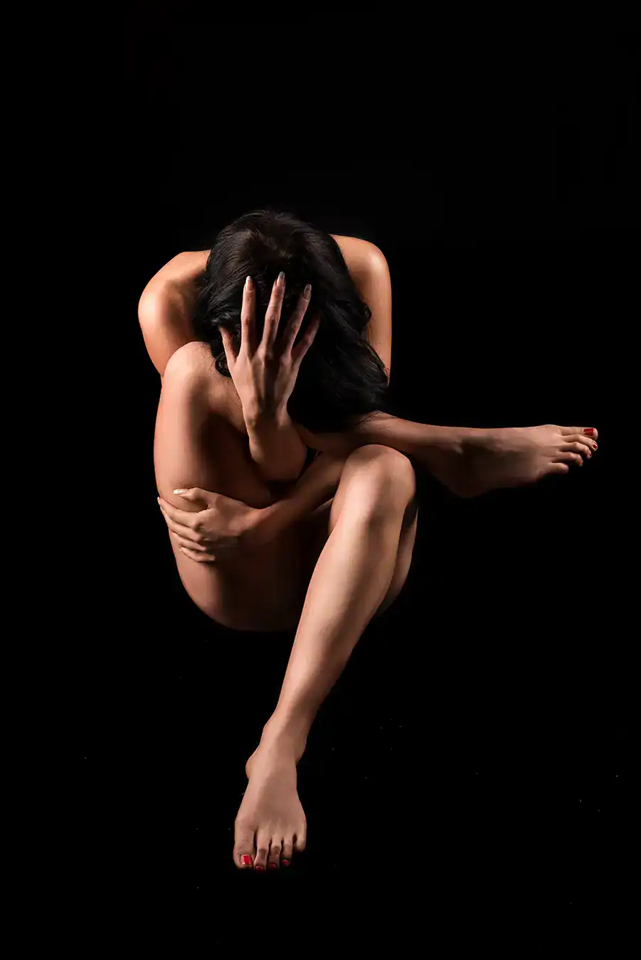 A captivating artistic nude photograph of a woman seated in an emotional pose, her hands clutching her head and arms wrapped around her body. The image is captured in stark contrast against a dark background, emphasizing the fluidity of the human form and the intensity of the emotion conveyed. The model's graceful posture and expressive gaze highlight both vulnerability and strength, creating a powerful and intimate composition.