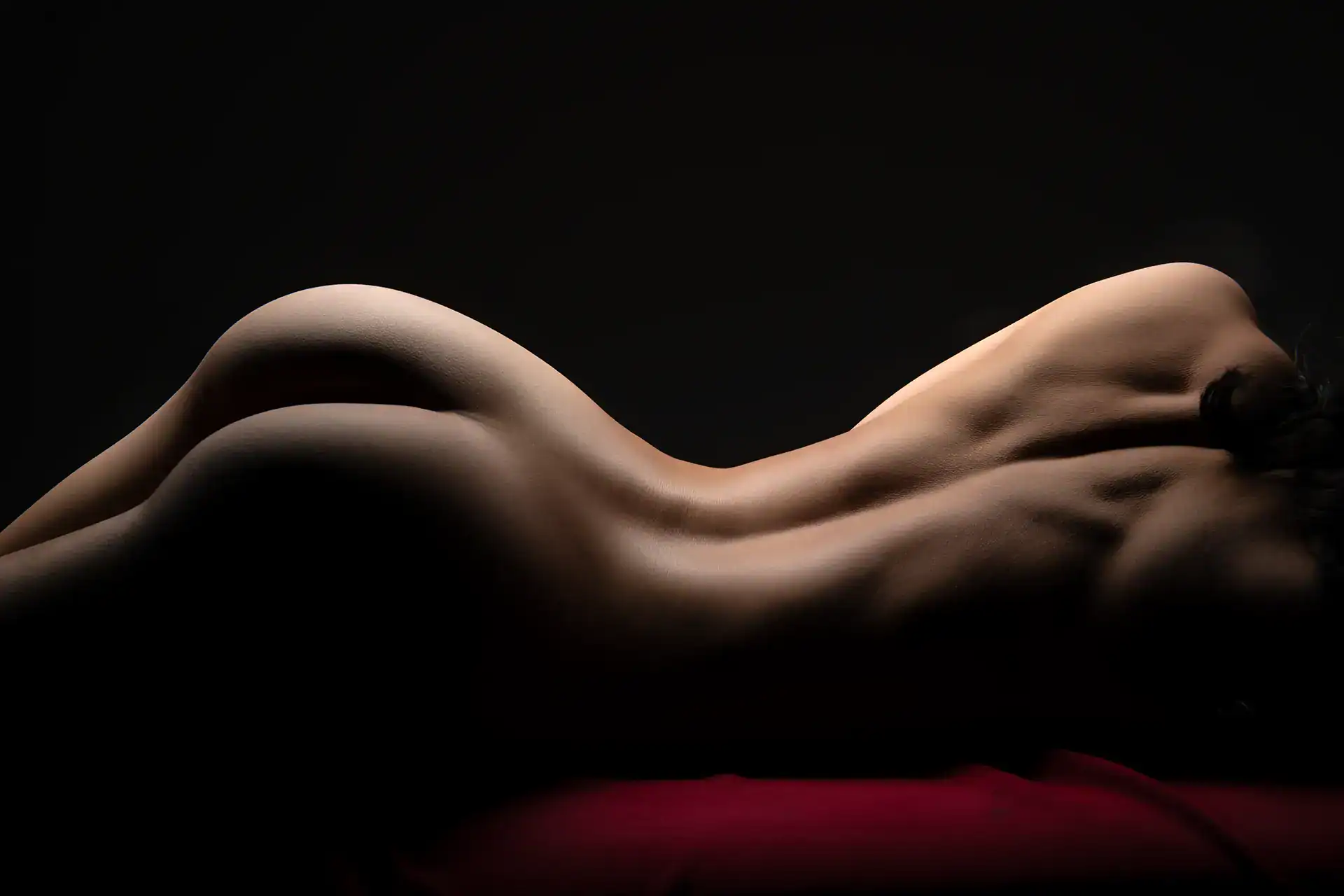 Minimalist bodyscape of a nude woman lying on her side, muscles and curves highlighted by soft lighting, set against a red fabric with dark background, Johannesburg artistic nude photography
