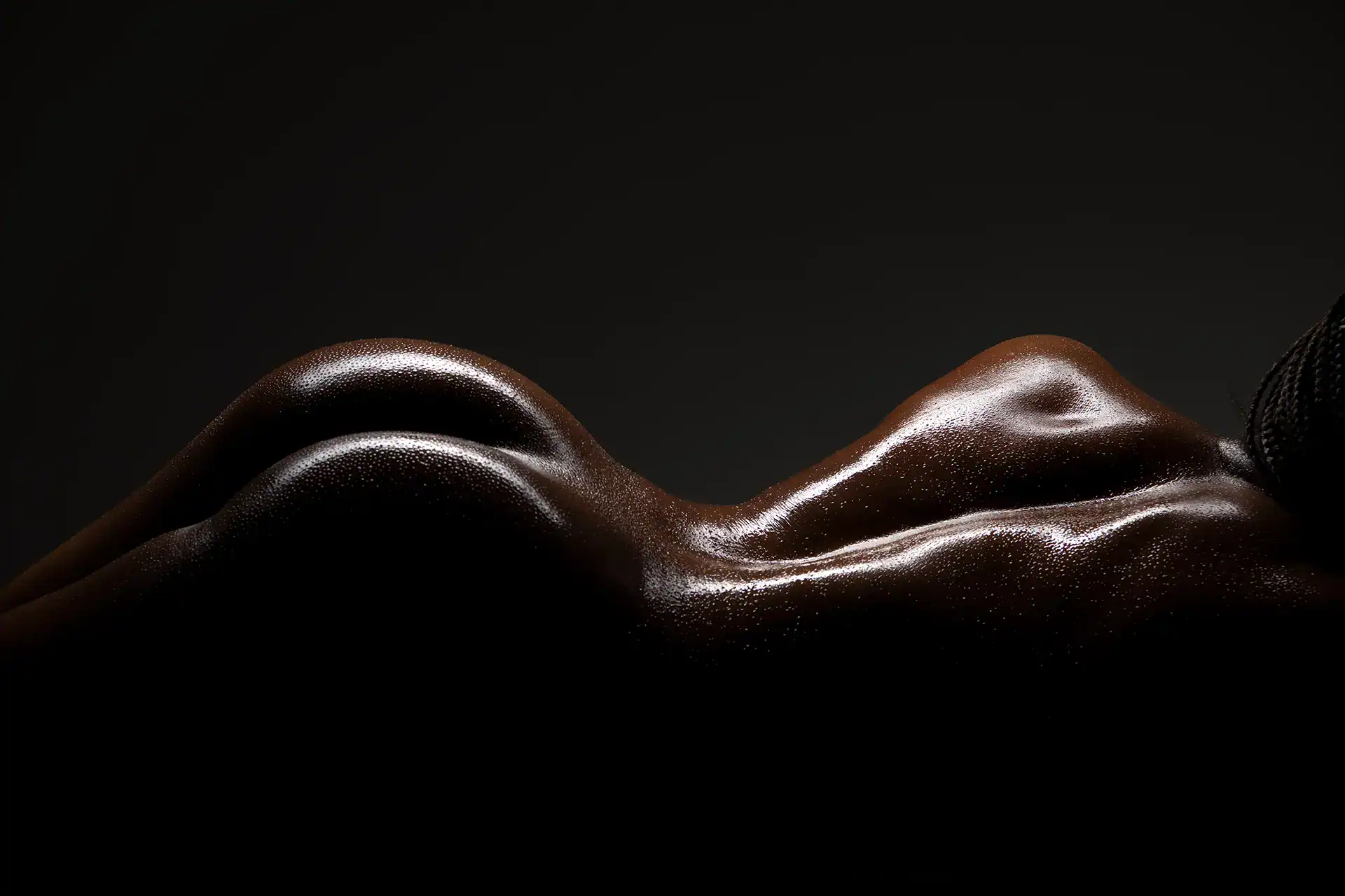 Close-up nude of a Black woman’s back and hips, glistening with moisture and highlighted by directional light, celebrating natural curves and skin texture in a premium African-inspired fine art nude session