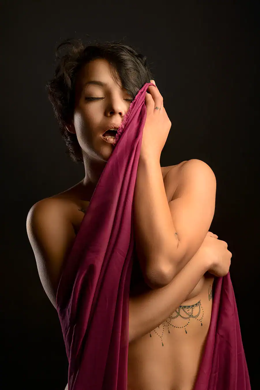 Close-up boudoir portrait of a topless woman embracing herself with a crimson drape against a dark backdrop, eyes closed in sensual expression, captured at Boudoir Fusion Photography