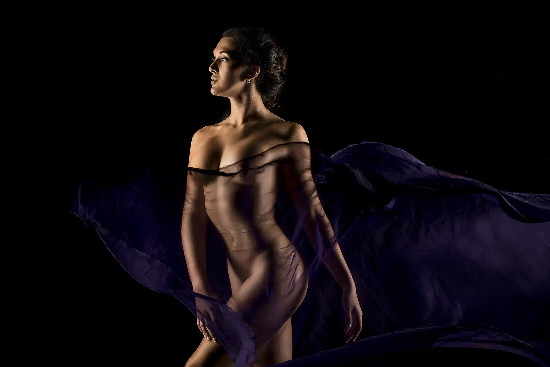 A dramatic nude photograph capturing the beauty of the female form, with flowing purple fabric artistically draped around the model’s body. The fabric moves gracefully, enhancing the sensuality of the pose, while the dark background creates a high-contrast effect, drawing attention to the curves and details of the body. The model’s strong yet elegant posture highlights both strength and femininity in a dynamic, graceful composition.