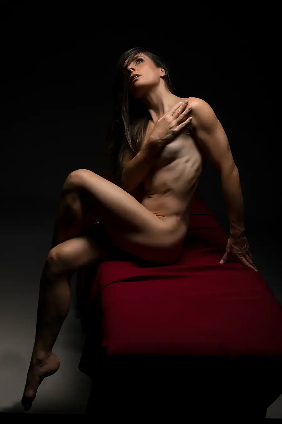 A striking nude portrait of a woman in a powerful pose, captured with strong lighting that highlights her muscular physique and graceful curves. The deep shadows emphasize her strong yet elegant form as she rests on a red fabric backdrop, exuding confidence and strength in the composition.
