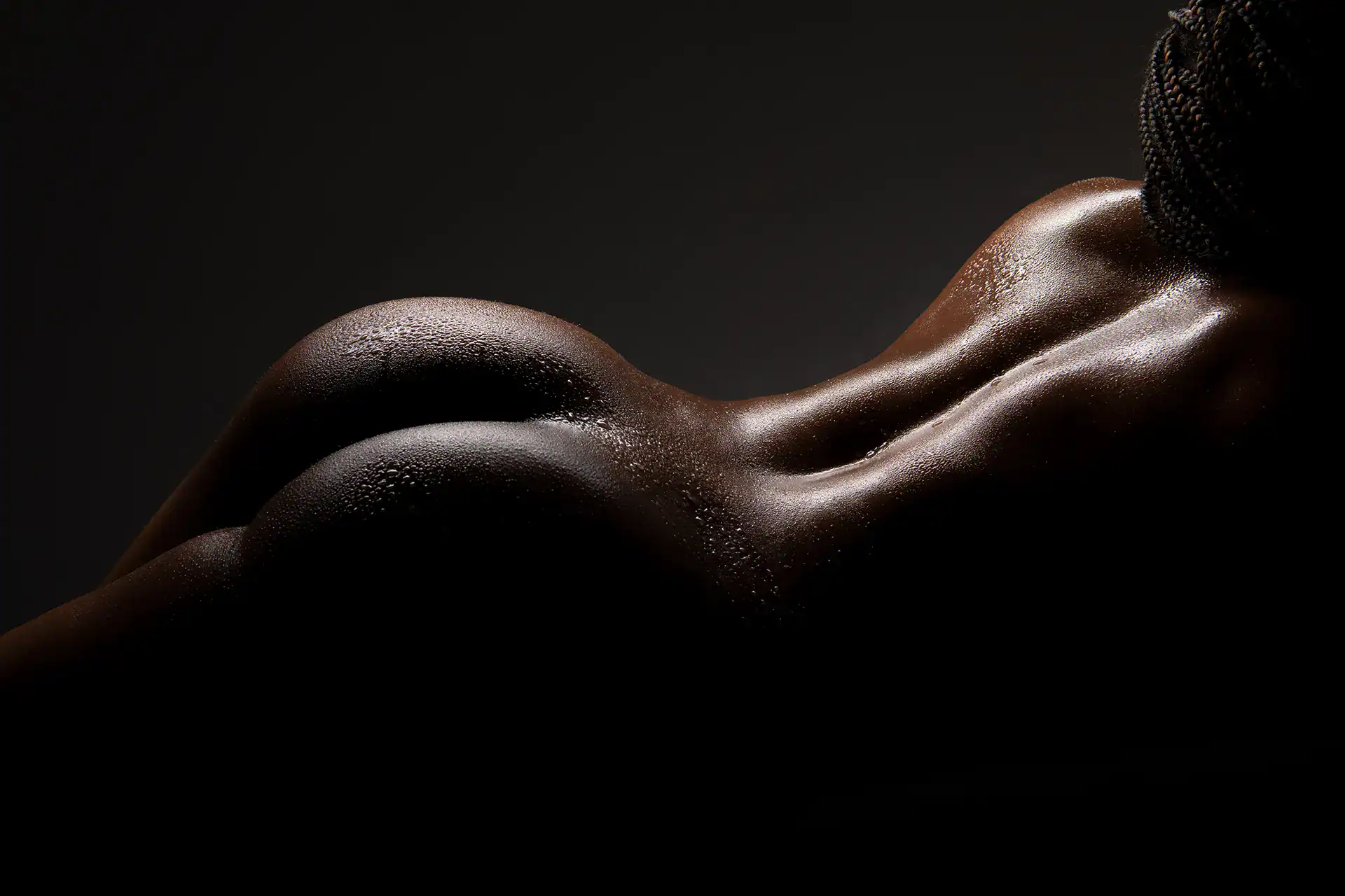 Rear nude view of a Black woman with braided hair, water droplets glistening along her back and hips under soft studio lighting, captured in an empowering fine art nude session in Johannesburg