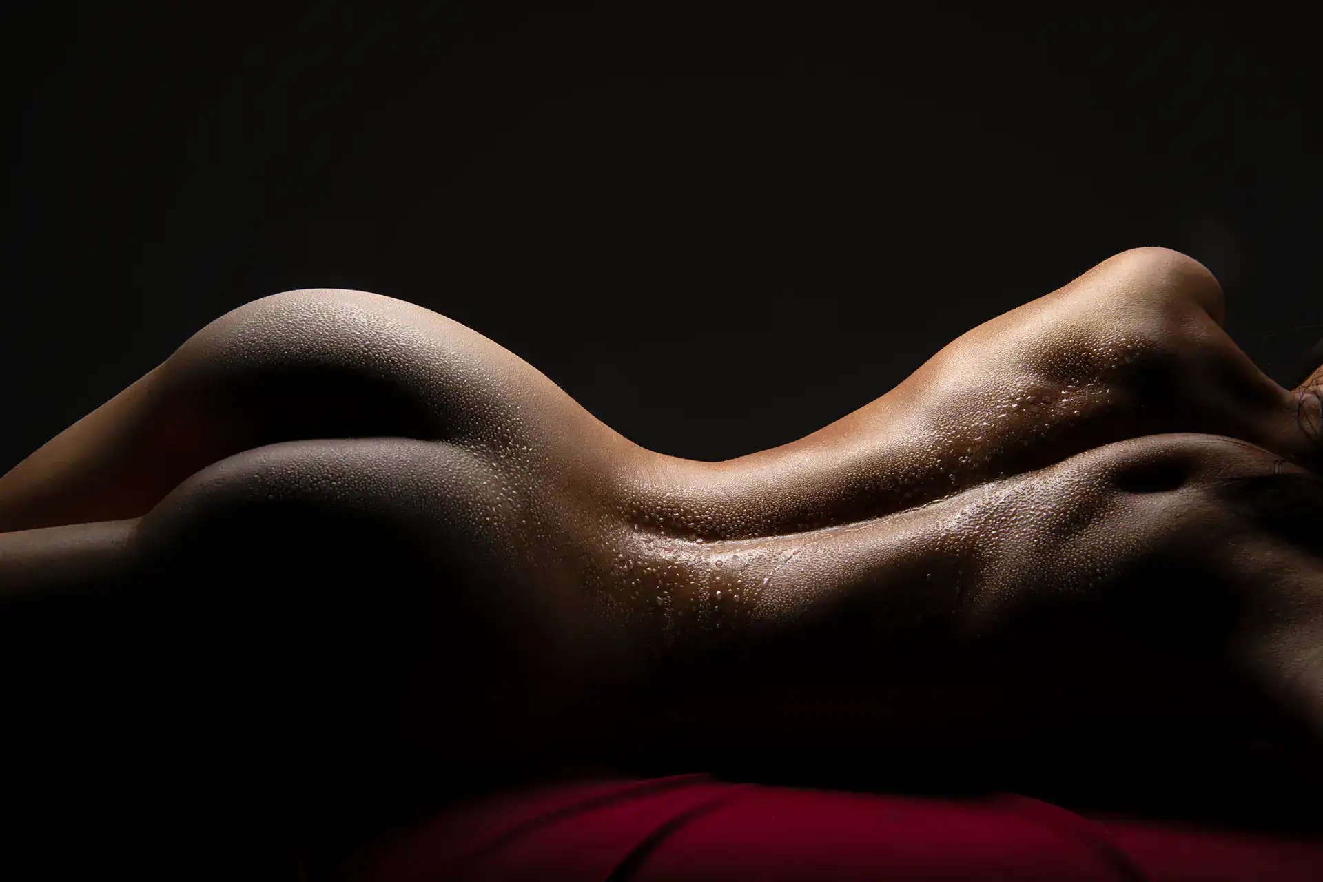 Sensual nude figure captured in profile with water droplets glistening on the skin, reclining on a red drape under soft low-key lighting, Johannesburg bodyscape photography