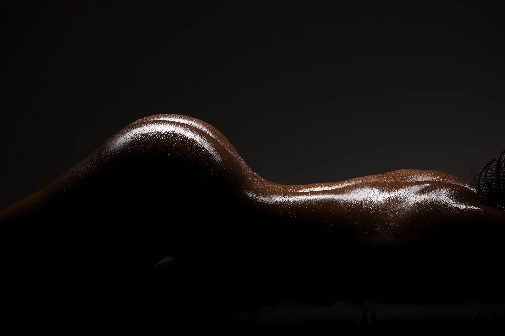 Fine art nude of a Black woman lying on her stomach, her glistening curves softly illuminated in studio light, showcasing rich skin tones and powerful femininity by a South African fine art nude specialist