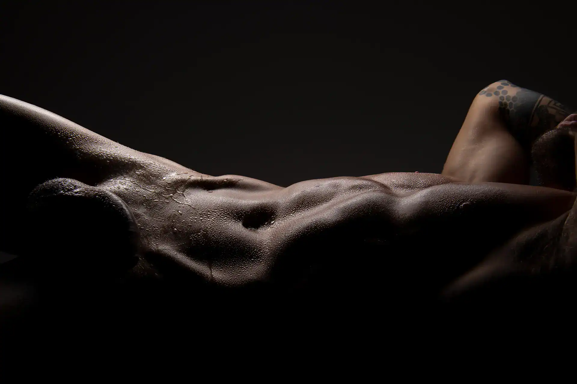 Artistic male nude featuring a glistening, muscular torso and tattooed arm, droplets catching the light in a low-key studio session with a male fine art nude photographer based in South Africa