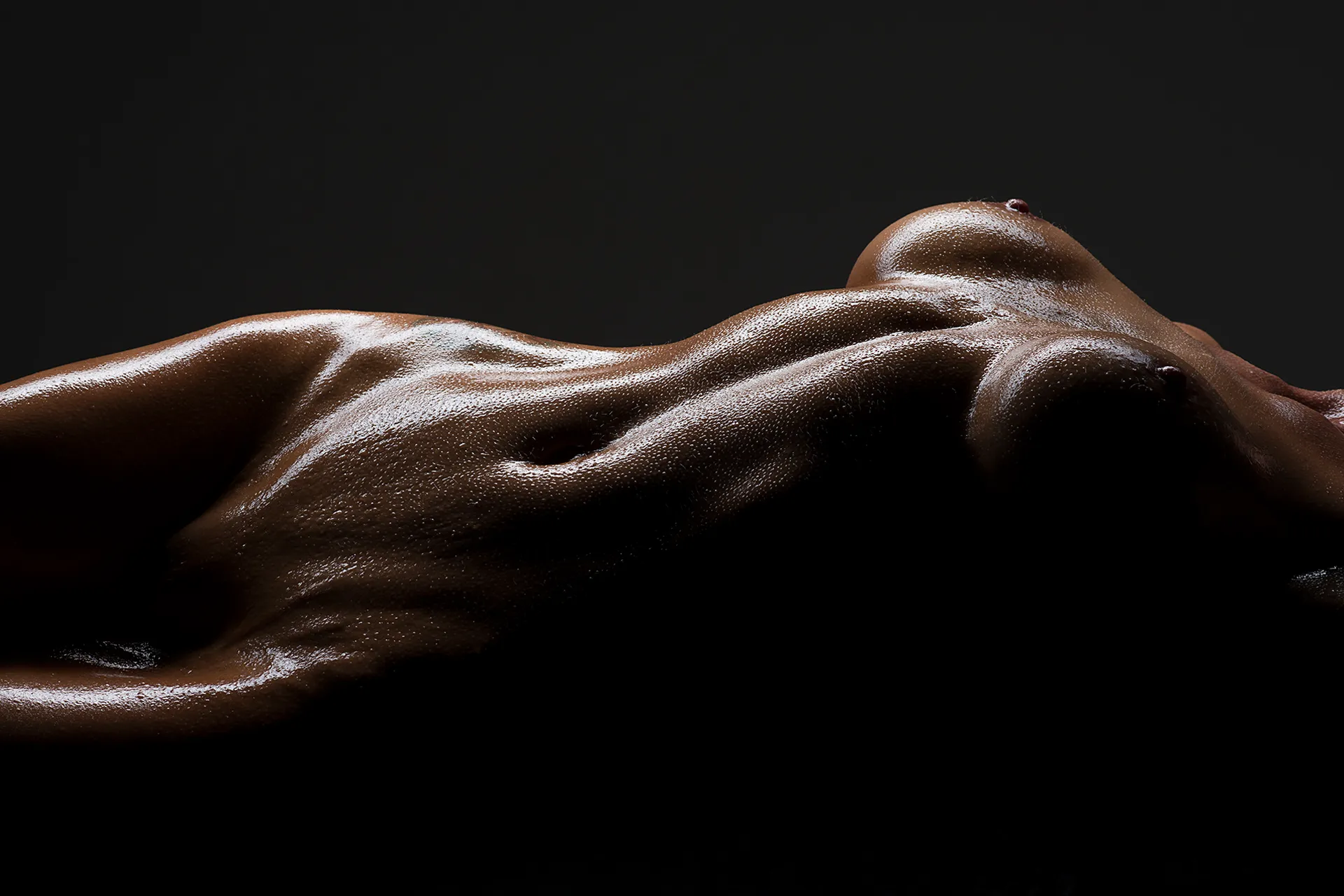 Bodyscape of a nude torso lying on back, covered in oil and water droplets, captured in moody dramatic lighting at Boudoir Fusion Photography in Johannesburg