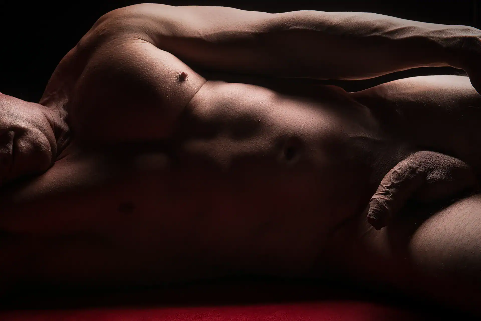Fine art nude of a male model lying on his side on a bed with red velvet, uncut flaccid penis visible, captured by an erotic photographer in Johannesburg, celebrating male sensuality and softness through artistic composition