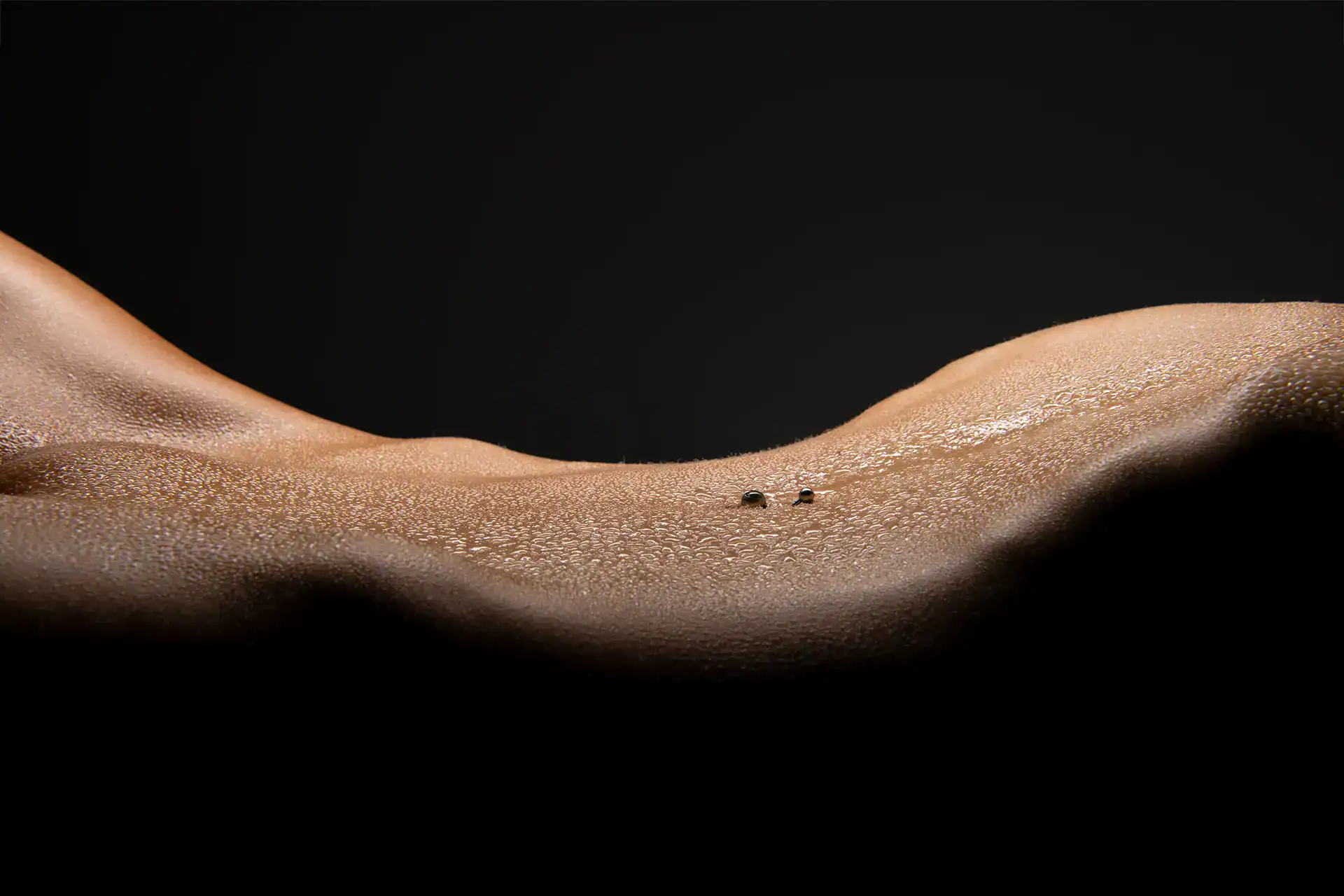 Close-up of a glistening nude torso with water droplets, highlighting the curves of a pierced navel under soft light, Johannesburg fine art bodyscape photography