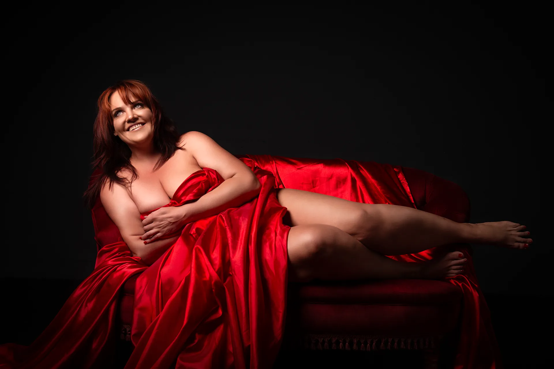 Beautiful boudoir photography of a red-haired woman lying on her side, draped in a flowing red silk sheet on a matching red couch, creating a passionate and elegant scene