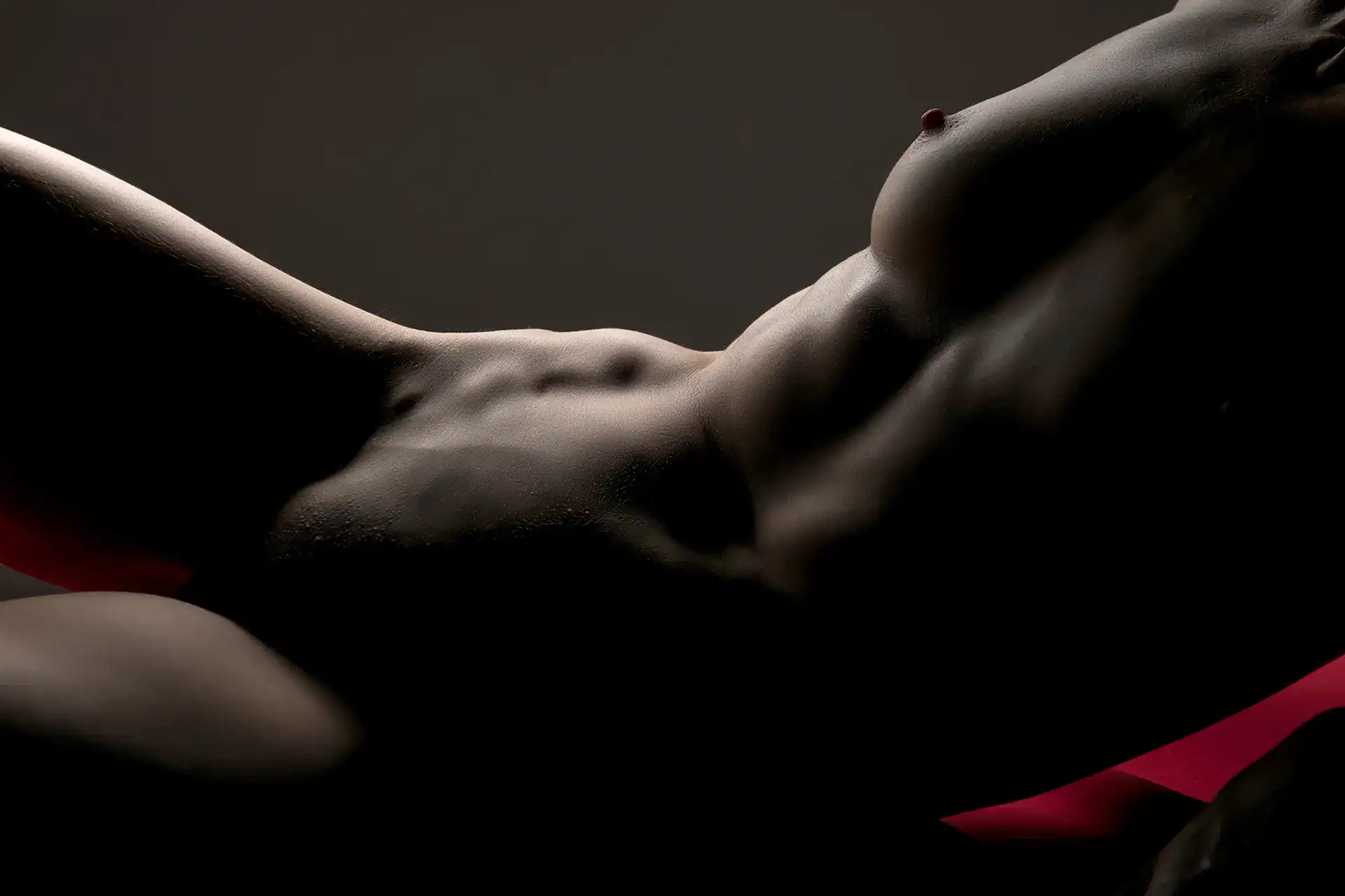 Side-lit artistic nude of a woman reclining with one breast exposed, capturing muscle definition and dramatic contrast, set against a soft red cloth, Johannesburg nude photography