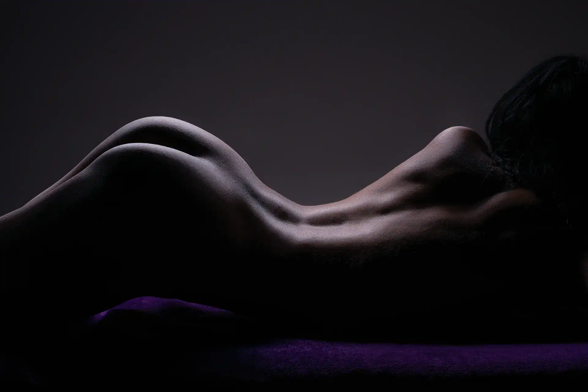 Artistic nude of a woman lying on her side, lit to highlight the curve of her back and shoulders, against a moody purple-toned backdrop, Johannesburg fine art bodyscape