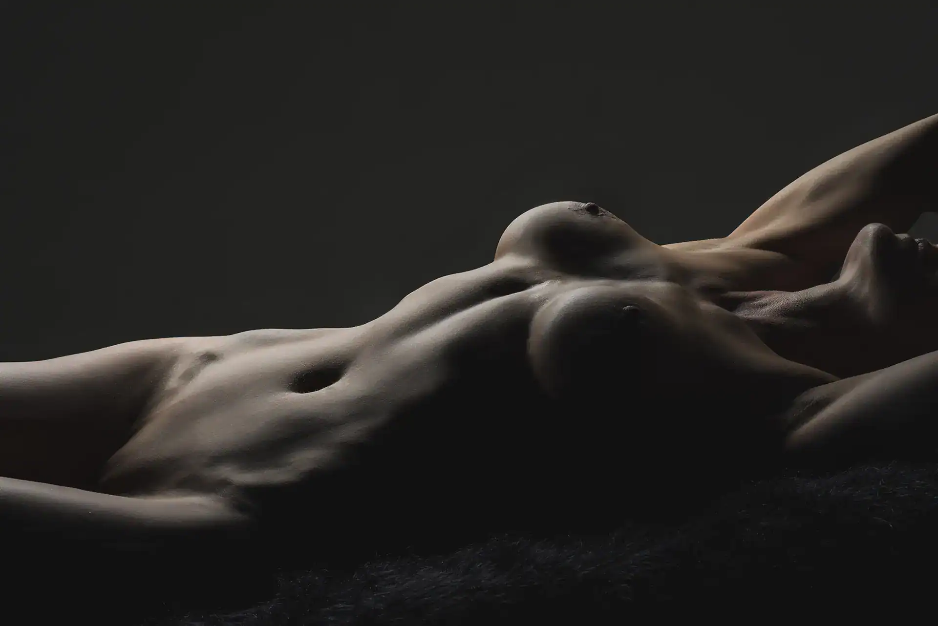 High-contrast nude of a reclining woman with visible breasts and defined abs, partially lit face and arms adding emotion and depth, captured in Johannesburg studio nude photography